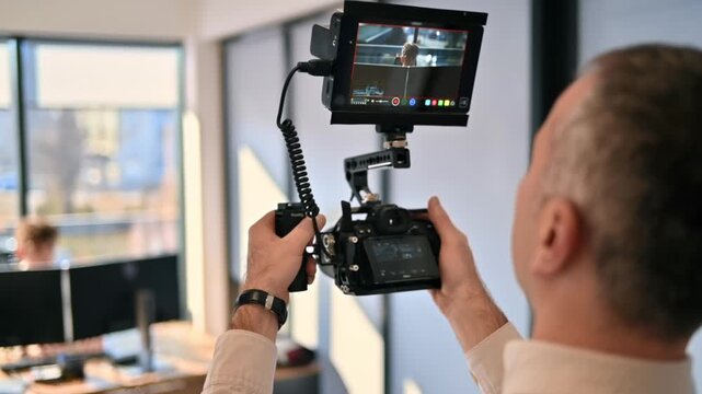 Cameraman shooting video footage in office setting, using professional camera with external monitor for dynamic recording.