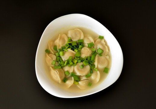 A deep plate of freshly cooked dumplings with broth and chopped green onions against a black background.