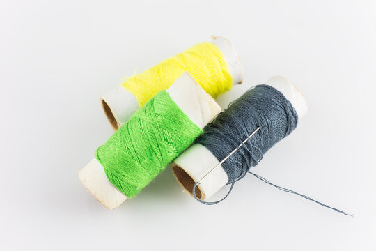Three colored thread spools and a sewing needle arranged on a plain white background, for sewing or craft use.