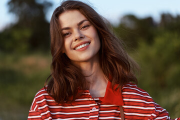 Fototapeta premium A smiling young woman outdoors wears a red striped polo, enjoying a sunny day. A lifestyle portrait emphasizing casual confidence, warmth, and natural beauty.
