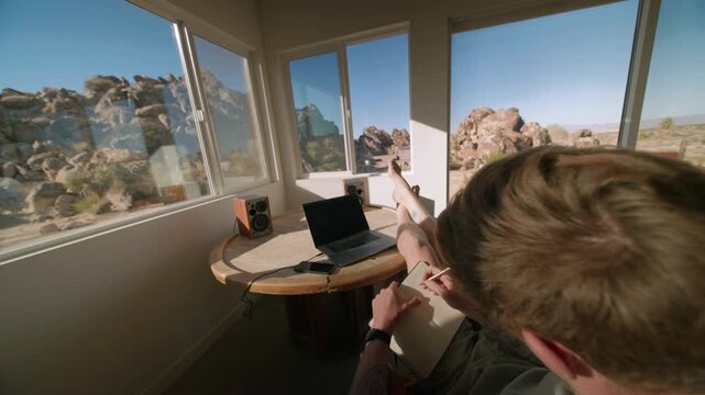 A musician reaches for his guitar beside a laptop at a round table with a rock formation view.