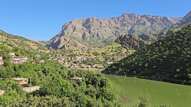 Emerald river winds past a mountain village nestled among lush hillsides, rugged peaks rising beneath a clear blue sky.
📍Selin village, Hawraman Takht, Kurdistan 