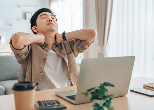 Young Asian man having neck and shoulder pain after working on a laptop for too long. He is massaging his shoulder to try to relieve the pain. Office syndrome concept