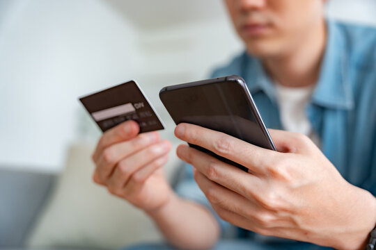 Close-up image of a man's hand holding a credit card and using a smartphone to make online purchases via internet banking in his living room at home