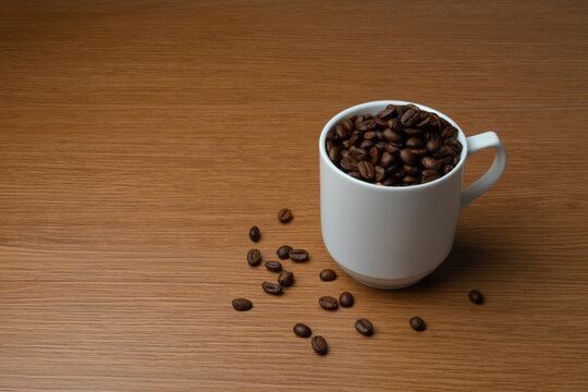 White mug filled with dark roasted coffee beans on a wooden tabletop with scattered beans. Copy space for text.