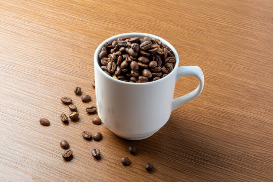 A white mug filled with roasted coffee beans on a wooden table with scattered beans. Copy space for text.
