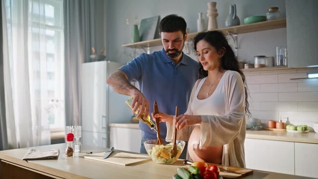 Married couple cooking salad kitchen. Husband adds oil pregnant wife mixing.