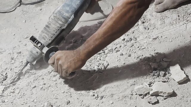 a construction worker using an electric rotary hammer drill to break up a concrete floor along a chalk line guide. The tool chips away at the hard surface, throwing up dust and small rock debris
