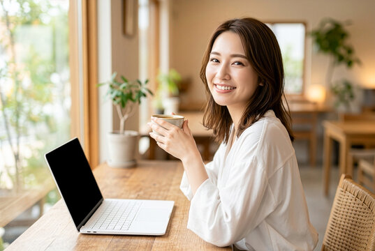 カフェでのリモートワーク風景｜コーヒーを手に笑顔を見せながらノートパソコンを使う日本人女性