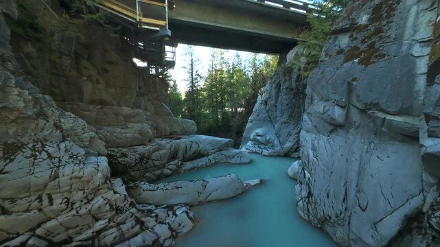 A descending aerial shot flies down past stone canyon cliffs, skimming right above a milky turquoise mountain river as it snakes around a rugged rocky bend in the summer woods.