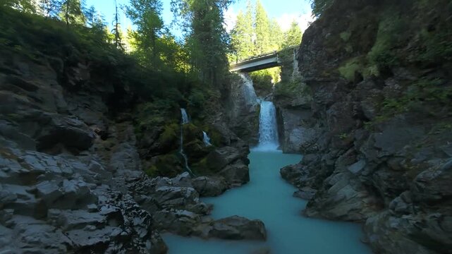 A slow forward tracking aerial shot glides gracefully through steep rock cliffs above a milky turquoise river pool toward a scenic cascading canyon waterfall during the summer.
