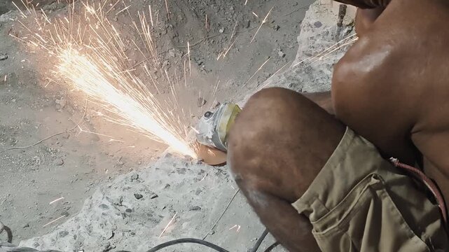 a construction worker using an electric angle grinder to cut through steel reinforcement bar embedded in a concrete structure. Bright orange sparks shower across the dusty concrete ground