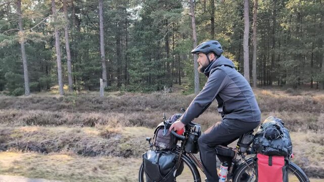 Adventurous man cycling on a touring bike loaded with gear for a bikepacking trip, riding along a paved path through a sunlit forest and heathland landscape