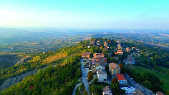 Picture-perfect aerial truck left shot of the green lands surrounding the Santa Vittoria in Matenano town center, full of thickets, ravines, buildings, roads, reaching distant misty hillscapes, clouds