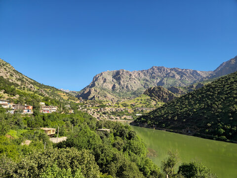 Emerald river winds past a mountain village nestled among lush hillsides, rugged peaks rising beneath a clear blue sky.
📍Selin village, Hawraman Takht, Kurdistan 