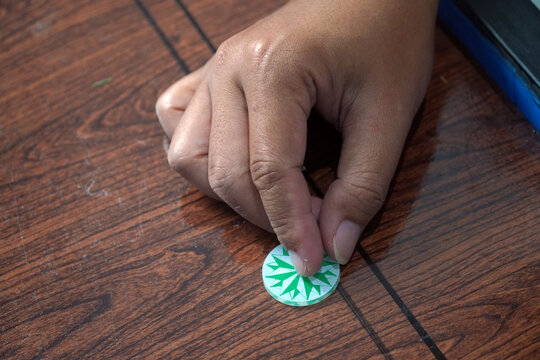 Close-up shot of a person's hand placing a green and white striker disc on a wooden carrom board surface. Focus on the finger and details of the plastic token. Traditional tabletop board game. karambo