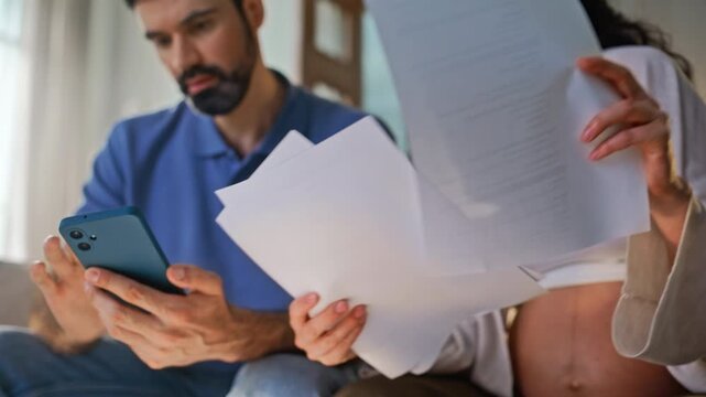 Worried couple checking bills home. Pregnant woman husband reviewing documents.