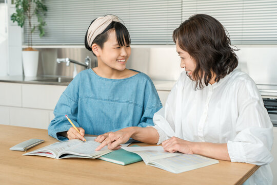 A girl is being tutored by her mother in the living room and kitchen of her home.