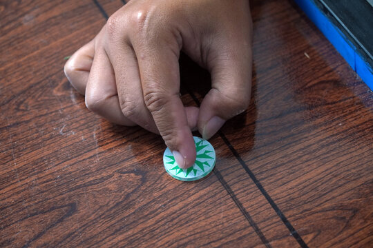 Close-up shot of a person's hand placing a green and white striker disc on a wooden carrom board surface. Focus on the finger and details of the plastic token. Traditional tabletop board game. karambo