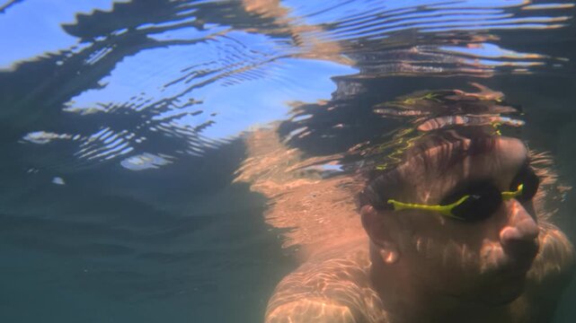 Man swimming underwater wearing yellow goggles, eyes closed, holding his breath, with clear water and sky above him