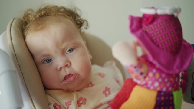 Baby girl with curly red hair sitting in a high chair, drooling, and looking at a colorful toy with a funny expression
