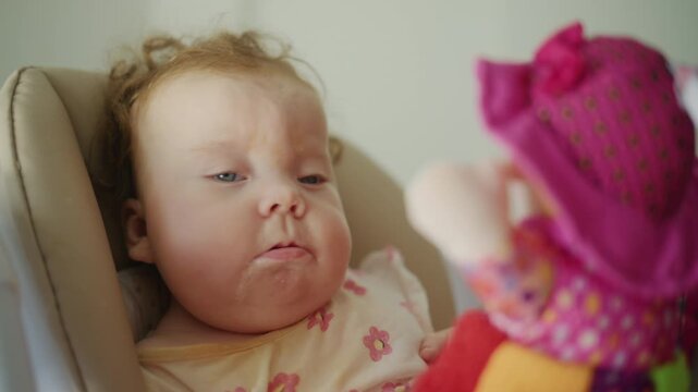 Baby girl sitting in highchair, drooling, and focusing on a brightly colored toy with curious eyes