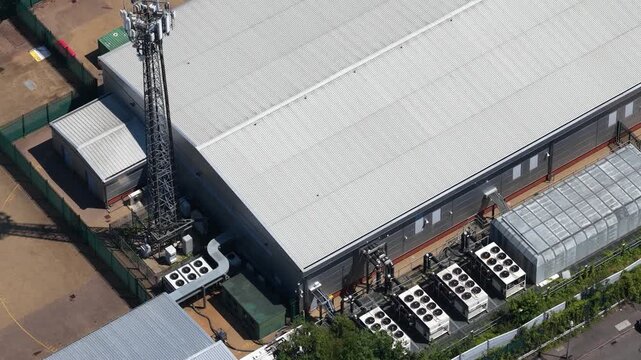 High angle drone shot showing a modern industrial warehouse with powerful air conditioning units for cooling, alongside a tall cellular communication tower for network connectivity