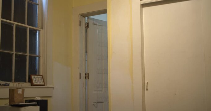 Still shot of a modest guest room interior with a window, open doorway to a lit room, and closed closet doors.