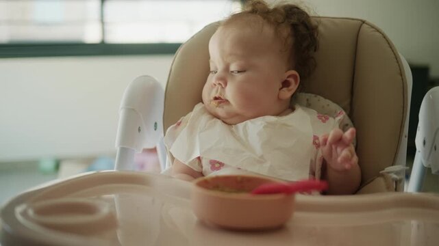 Baby falling asleep while eating a meal in a high chair at home, food smeared on bib and face during infancy