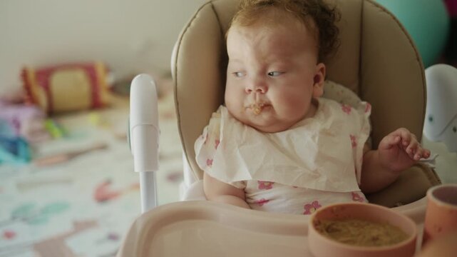 Young baby with a bib sitting in a high chair, being fed nutritious pureed food from a spoon by an adult hand