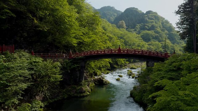 Iconic red Shinkyo Bridge spans the clear Daiya River amid lush green forest and mountains in Nikko, Japan, on a peaceful summer day.