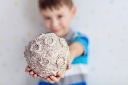 A young boy with a gentle smile holds a textured papier‑m&acirc;ch&eacute; moon model featuring realistic craters and muted gray tones; the soft studio lighting create a warm atmosphere.