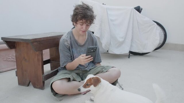 Teenager smiles and caresses an affectionate dog while briefly looking up from a smartphone after receiving good news. A positive interaction.