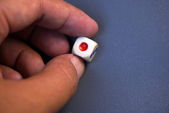 Close up of hand holding single white game die with red dot number one on blue background, luck, chance, board game, gamble, strategy, and decision concept.