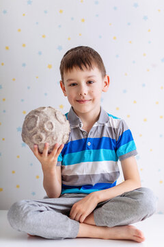 A cheerful young boy in a striped polo shirt and gray pants sits cross‑legged holding a textured papier‑m&acirc;ch&eacute; moon with realistic craters;the pastel star‑patterned backdrop create a playful atmosphere