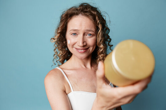 Authentic middle aged woman with natural skin applies cream from a jar. Close up on a soft blue background. Skincare, wellness, selfcare concept with untouched complexion and warm, inviting mood.