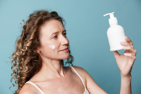 Middle-aged woman with curly hair looking at cream bottle. Natural skin without retouching. Skincare, wellness, and self-care concept on blue background studio.