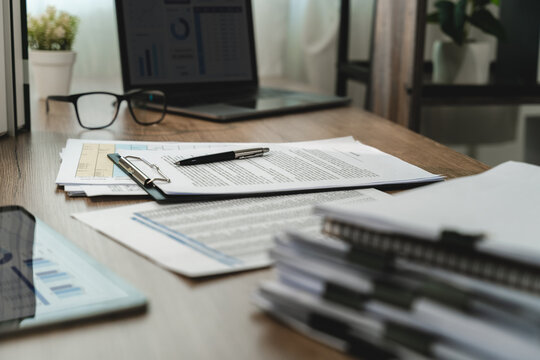 Printed business documents, clipboard, pen and eyeglasses arranged on an office desk beside a laptop, showing paperwork organization, report preparation and administrative workflow.