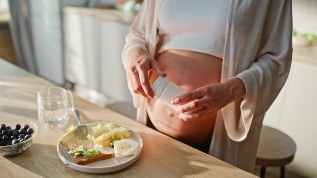 Pregnant woman taking prenatal vitamin pill kitchen. Close up belly supplement.