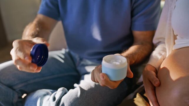 Man applying cream pregnant wife belly. Closeup hands caring baby bump home.