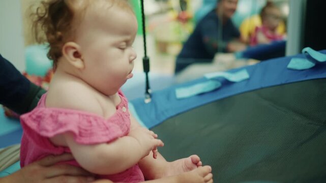 Little baby girl with a funny, grumpy expression sitting on an indoor trampoline, being held by an adult. Childhood lifestyle moment