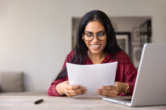 Indian woman in eyeglasses sits at table at homeoffice, reading documents with contented smile, feels satisfied with results, reviewing good business news. Success, achievement and professional growth