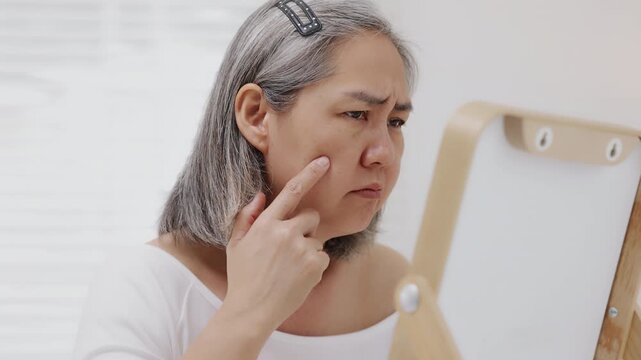 An Asian mature woman with grey hair looks in a mirror with concern while checking fine lines and aging skin.