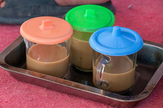 Three clear glasses containing a milk coffee or tea colored beverage, served on a metal tray with colorful plastic lids. A traditional style of serving coffee glasses on a metal tray.