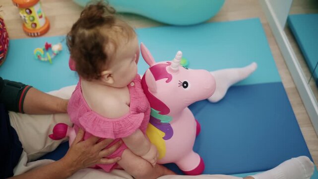 Baby girl playing and having fun with a pink unicorn bouncing toy on a blue mat, assisted by an adult