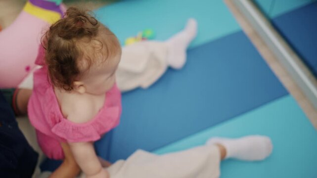 Baby girl in a pink dress sitting on a blue mat, playing indoors. Focusing on childhood and development