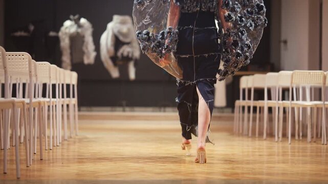 Fashion model walking on runway wearing unique designer dress with floral details and plastic elements during show.