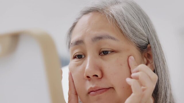 An Asian mature woman with grey hair looks in a mirror with concern while checking fine lines and aging skin.