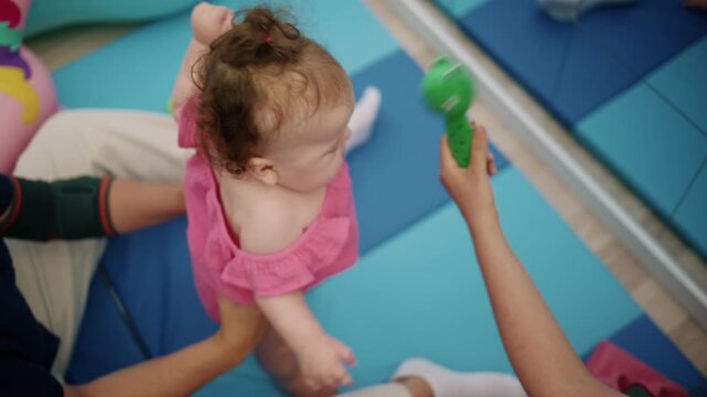 Baby girl in pink outfit learning to walk, parent holding her hands on a padded floor, developing motor skills