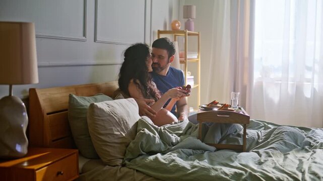Loving couple eating fruit bed cozy morning. Man embracing pregnant wife home.